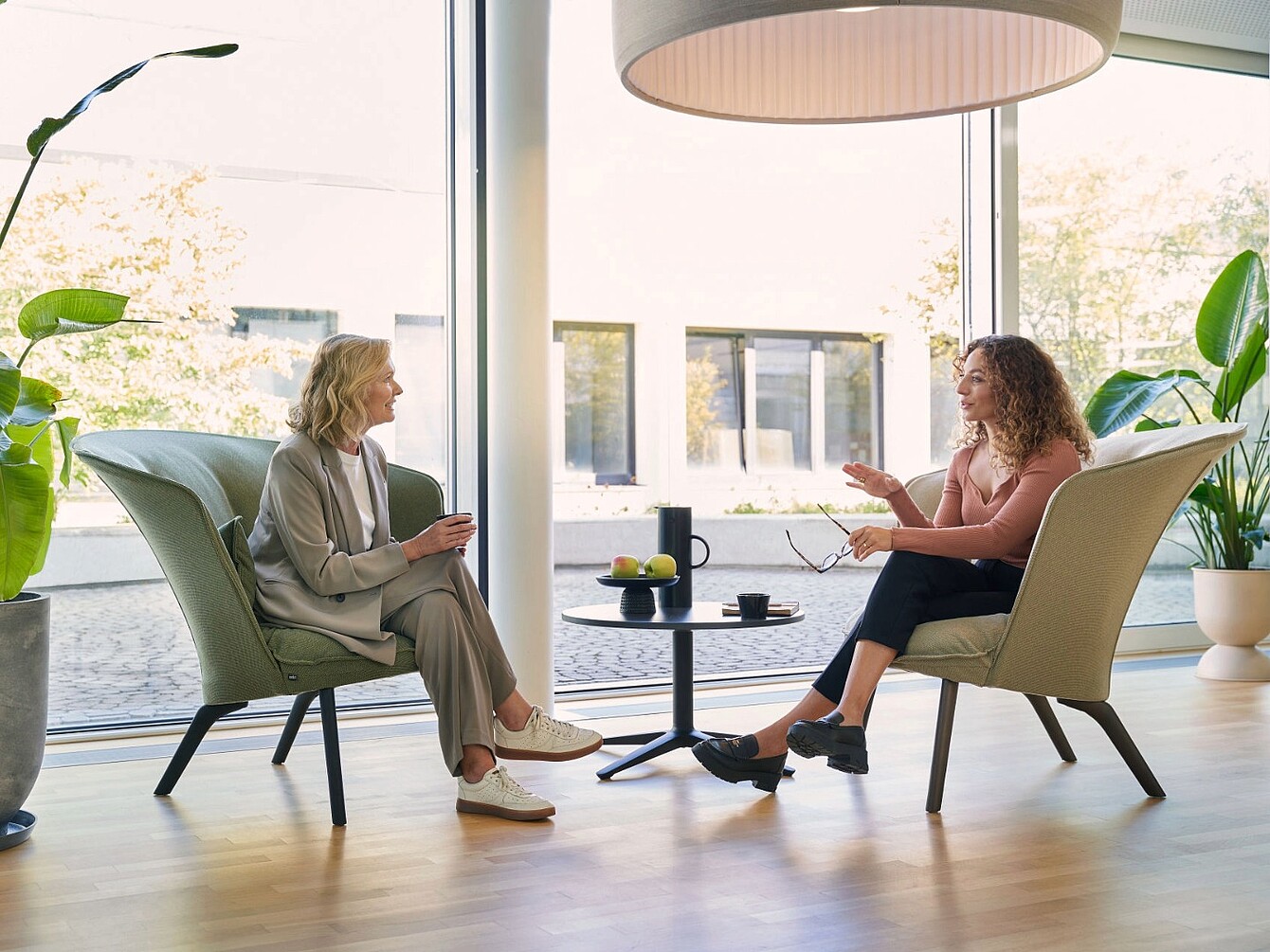 Two women sit opposite each other in modern armchairs having a conversation in a bright room with large windows and plants