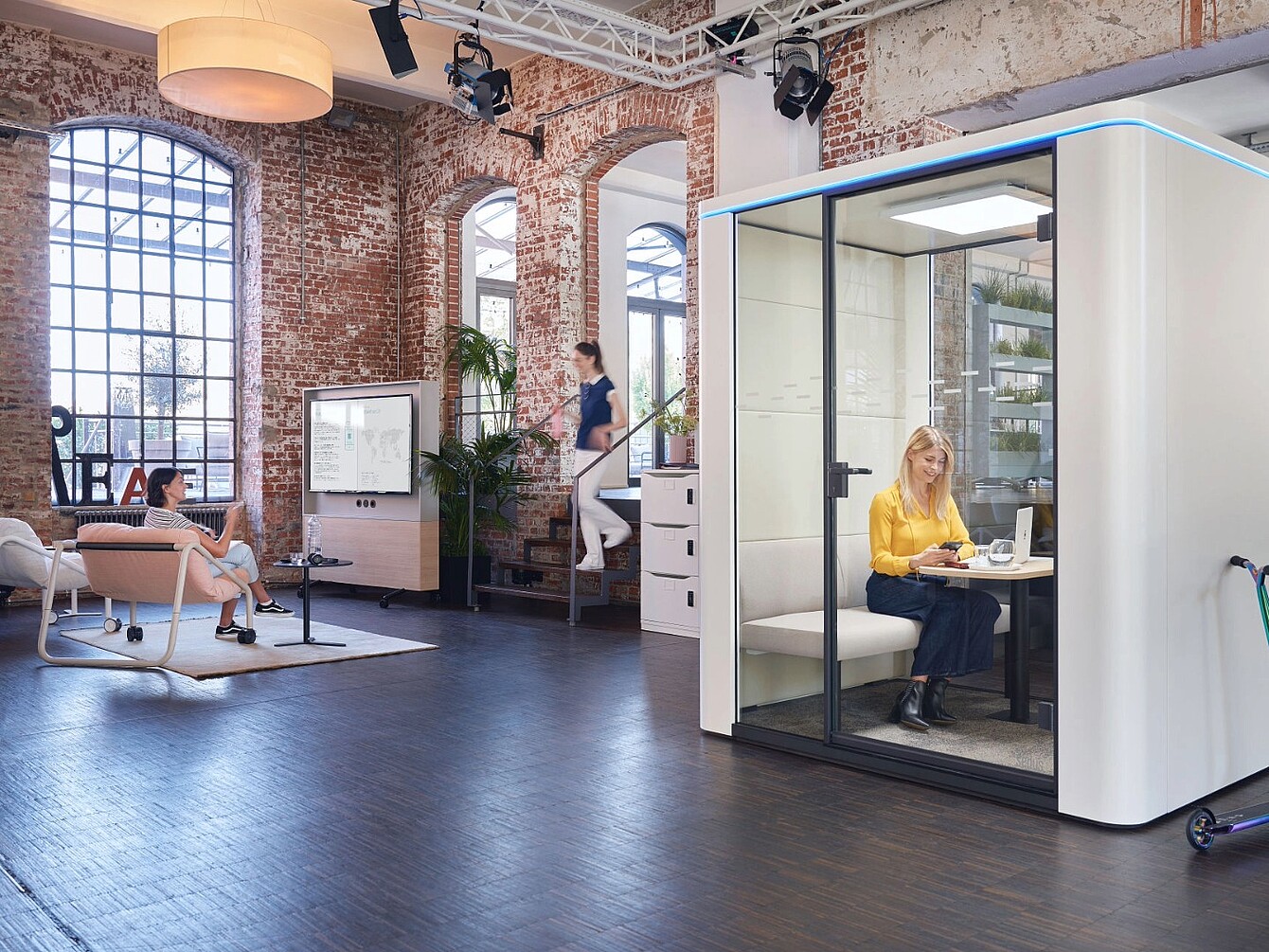 Offenes Büro mit Backsteinwänden, großen Fenstern, Sitzbereich mit zwei Personen und einer schallisolierten Telefonkabine, in der eine Frau sitzt
