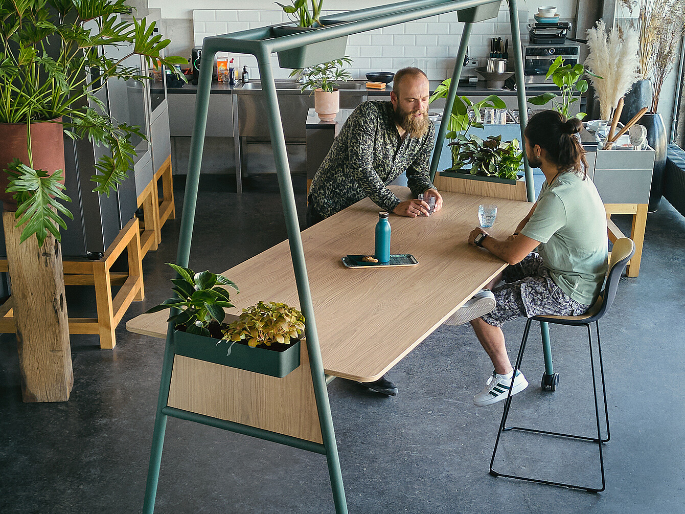 Two men conversing at a wooden table with integrated planters in a modern hybrid ductile commons workspace filled with plants and natural light