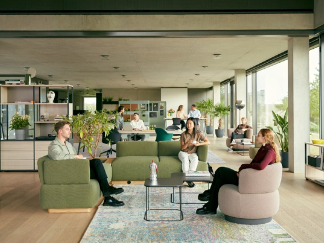 Several people sit on sofas and armchairs around a low table in a modern open office with large windows