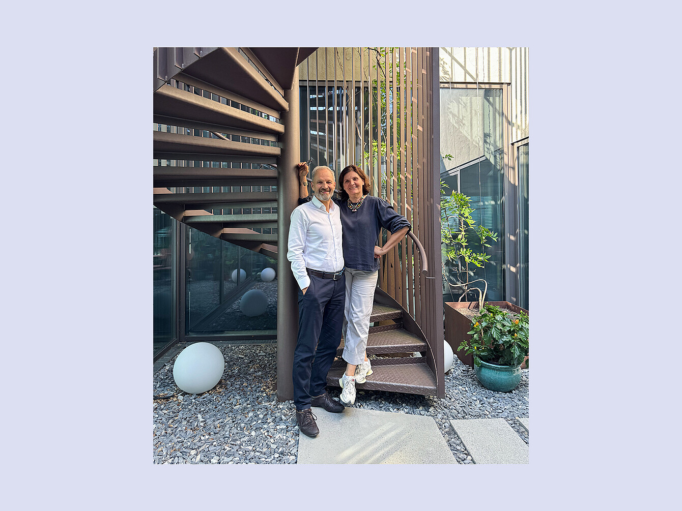 Zwei Personen (Helene und Markus Stainer) lehnen an einer spiralförmigen Außentreppe in einem Innenhof mit Kiesboden und Pflanzen in Töpfen