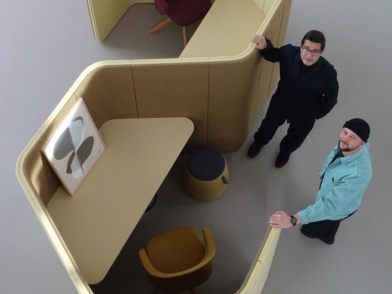 Zwei Männer (Rudi Raschke und Konstantin Thomas) stehen in einer modernen, beige-farbenen Arbeitskabine mit abgerundeten Trennwänden, einem Tisch, einem Hocker und einem Stuhl, auf dem ein gerahmtes Bild mit abstraktem Design lehnt.