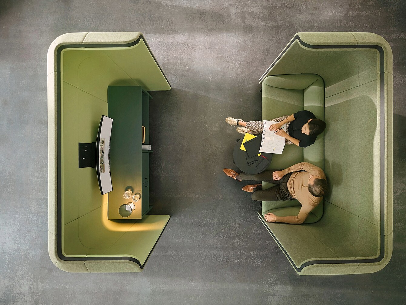 Zwei Personen sitzen in einem modernen, offenen Büroarbeitsplatz mit hohen, gepolsterten Trennwänden und einem kleinen Tisch zwischen ihnen, daneben ein weiterer Arbeitsplatz mit Bildschirm und Ablagefläche