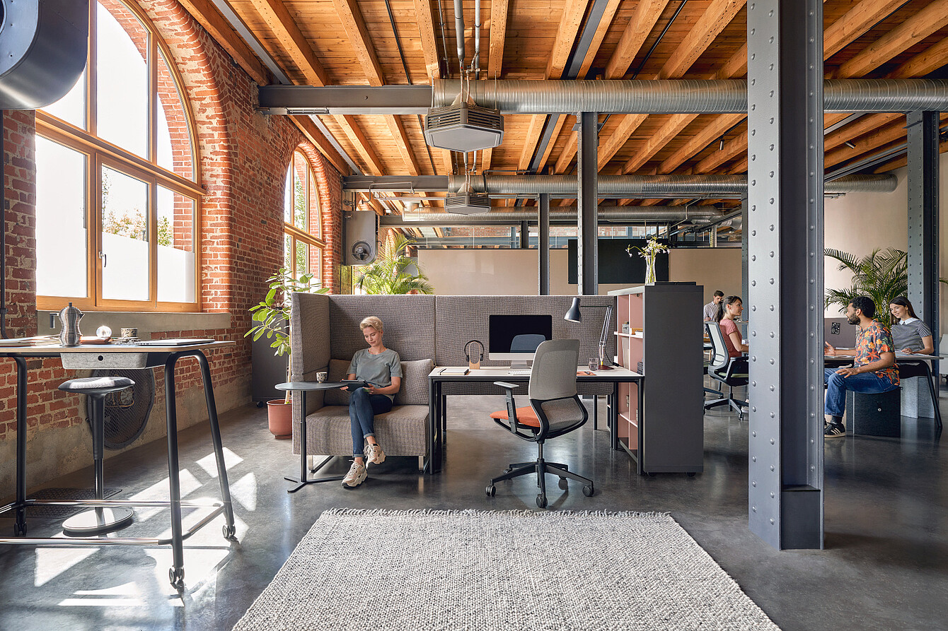 Open-plan office with exposed wooden ceiling beams, large arched windows, and metal support columns, featuring a person sitting on a high-backed sofa working on a laptop and two people conversing at a table in the background