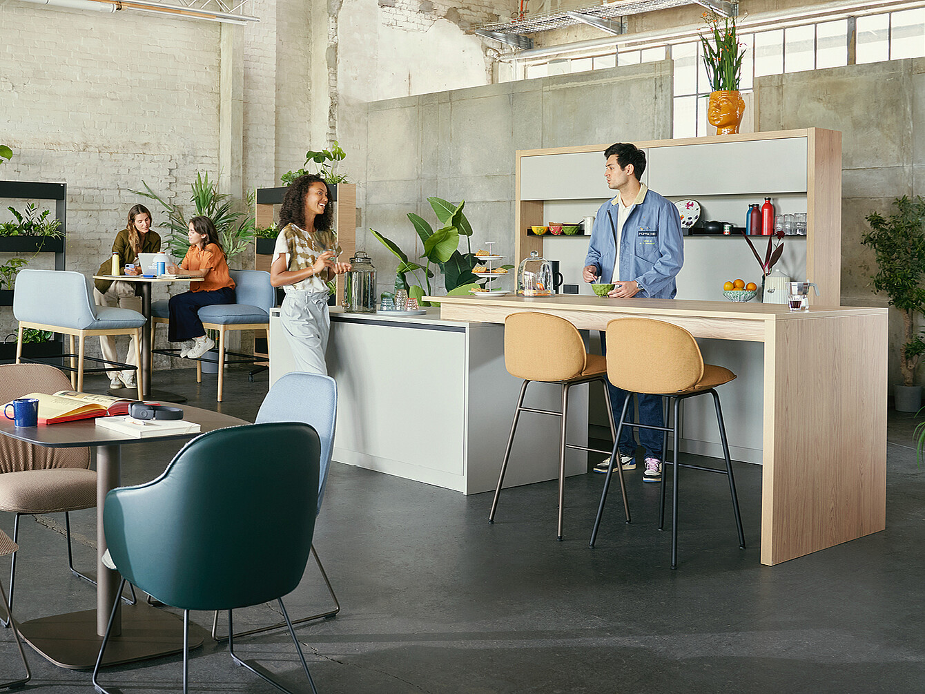Modern work café with flexible seating and a central counter where a man in a blue jacket serves a woman in a white apron, surrounded by plants and industrial-style walls