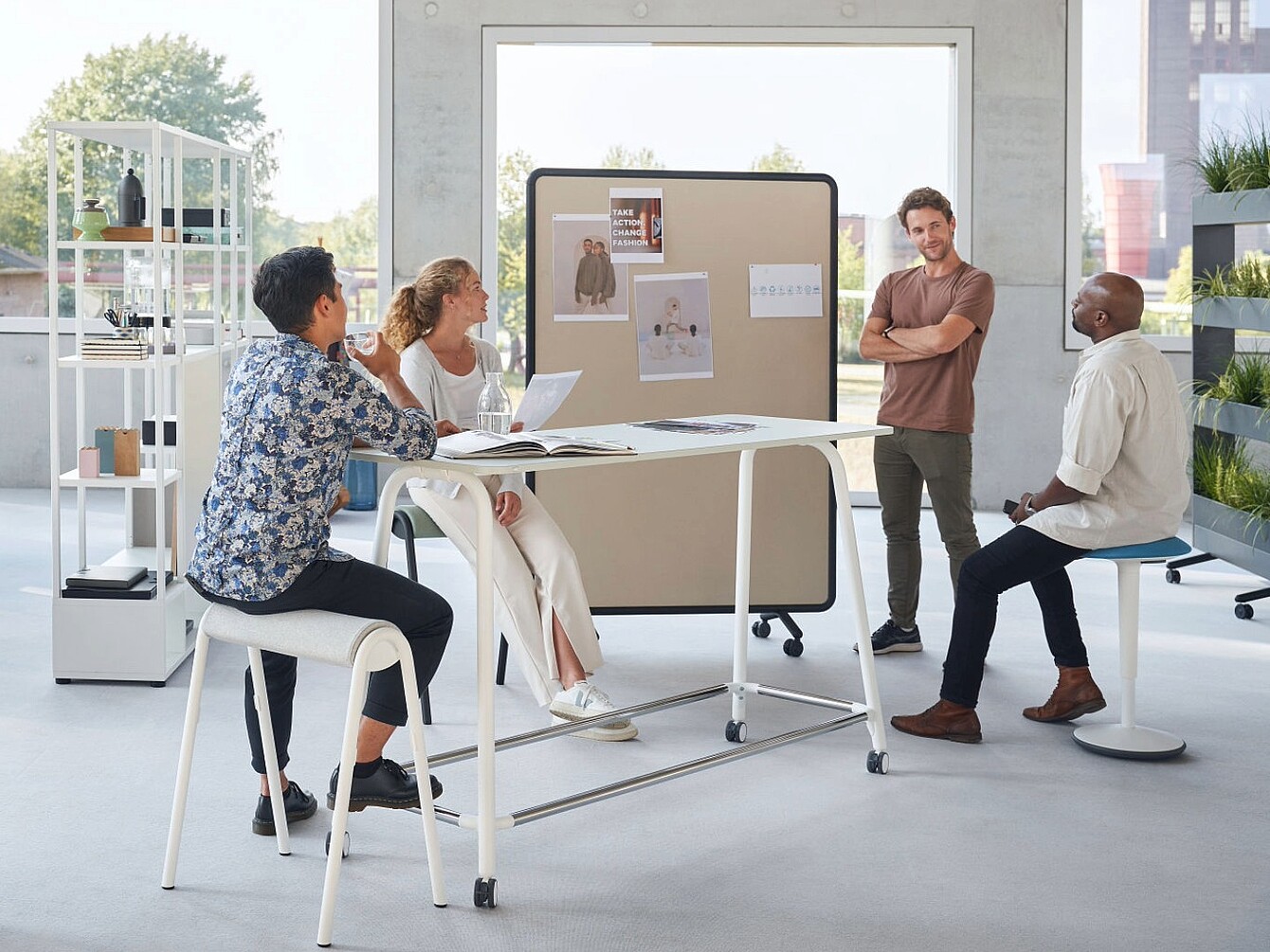 Das Bild zeigt einen modernen, offenen Bürobereich, in dem mehrere Personen an einem hohen Tisch zusammenarbeiten und Dokumente besprechen. Mobile Trenn-wände, Regale und Pflanzen strukturieren den Raum und schaffen flexible Zonen für Teamarbeit und Austausch. Die großen Fenster sorgen für viel Tageslicht und unter-stützen eine helle, freundliche Arbeitsumgebung.