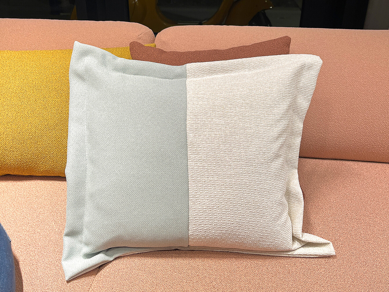 Zweifarbiges Kissen mit hellgrauer und cremefarbener Seite auf einem rosafarbenen Sofa, daneben ein gelbes und ein braunes Kissen.