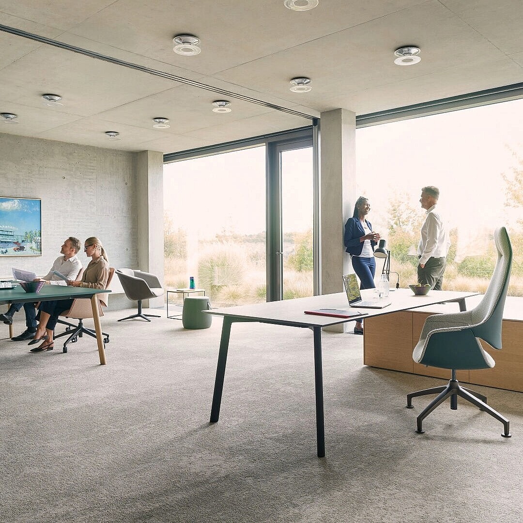 Modern office space with groups of people working and conversing near large windows and minimalist furniture