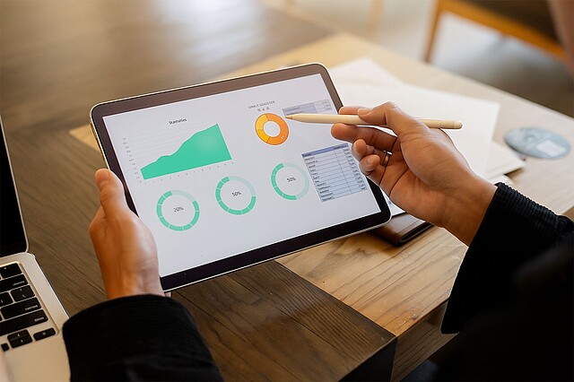 Person hält Tablet mit Diagrammen und Kreisdiagrammen zur Datenanalyse, zeigt mit Stift auf orangefarbenen Ring