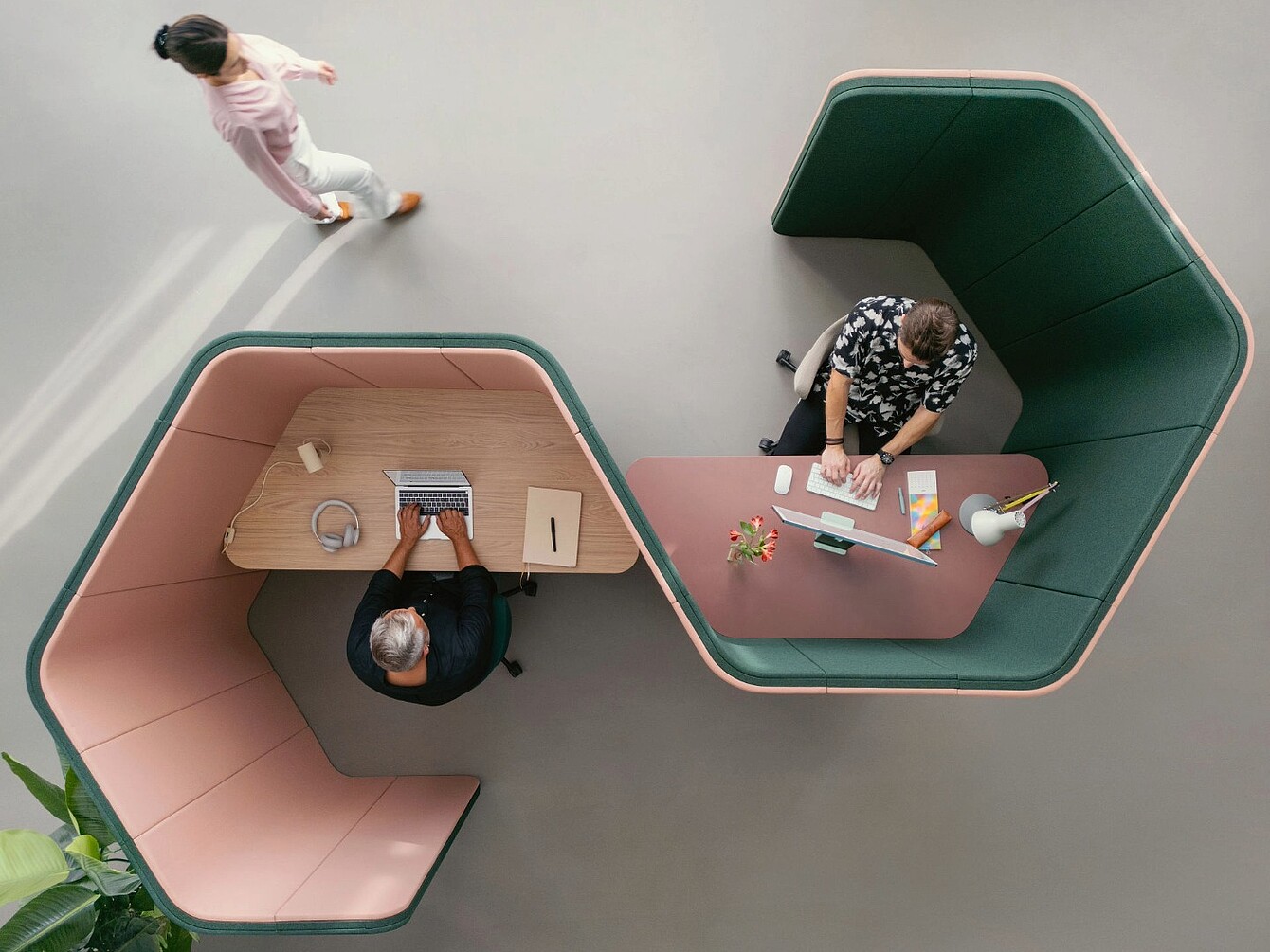 Zwei Personen sitzen an gegenüberliegenden se:hive-Raumlösungen mit akustisch gedämpften, geschwungenen Trennwänden, eine Person geht am Arbeitsplatz vorbei.