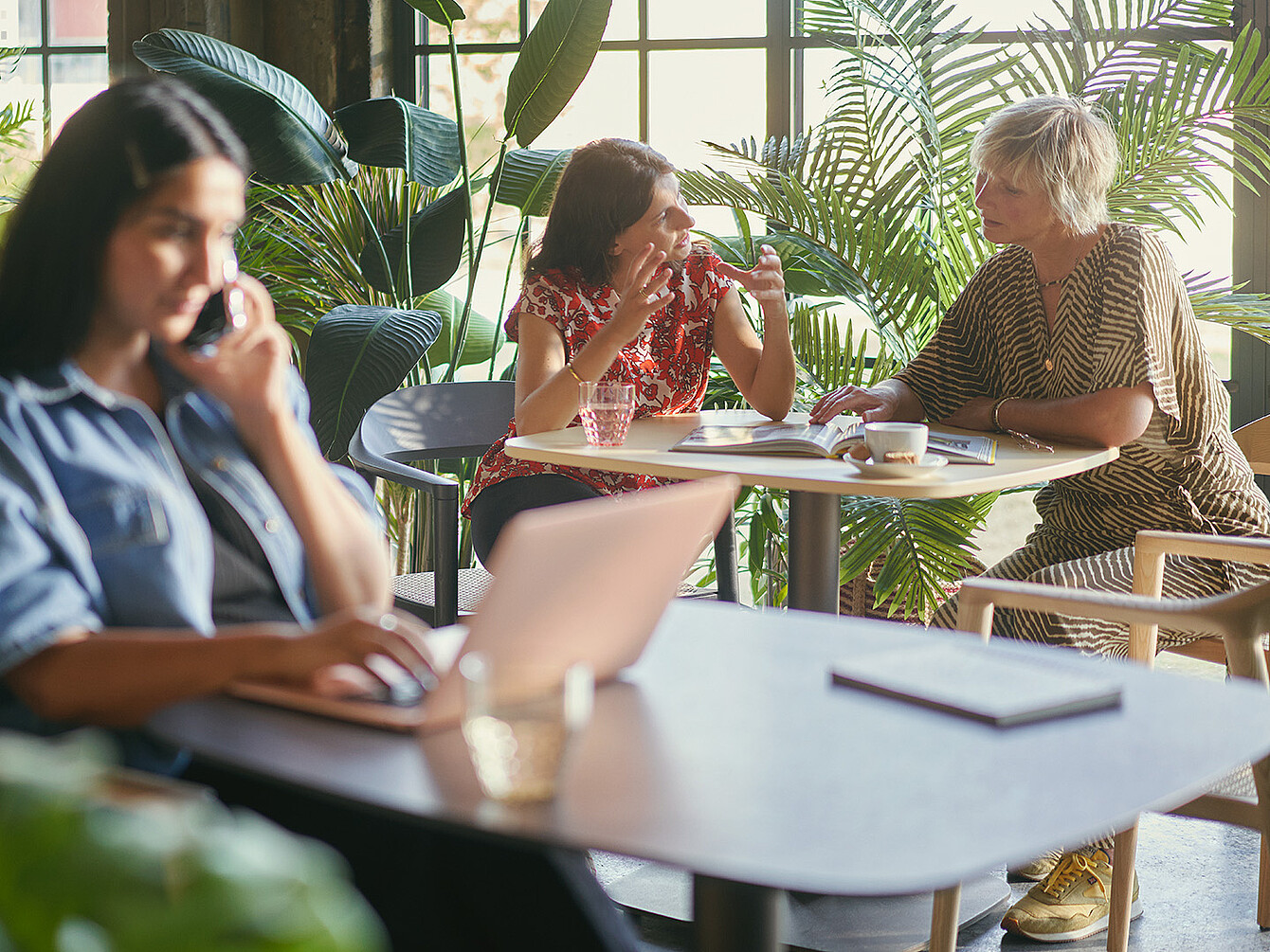 Three women in a bright Smart Café office space with large windows and plants; one works on a laptop while two others discuss over documents at a table