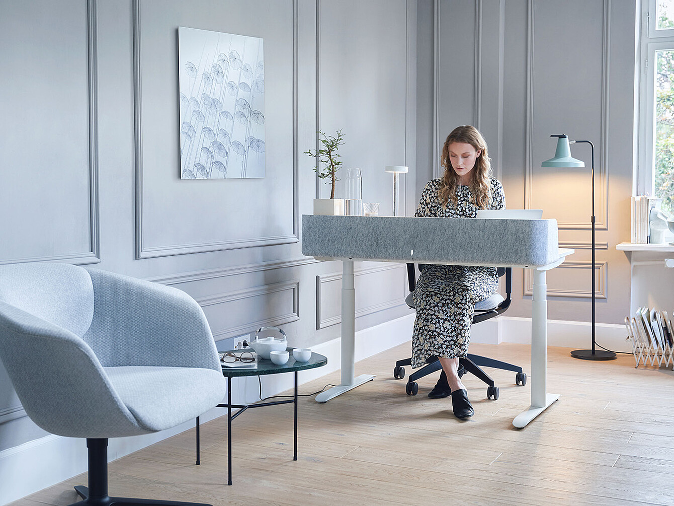 Woman in floral dress sitting at a modern white Sedus desk in a light gray room with a blue armchair and floor lamp