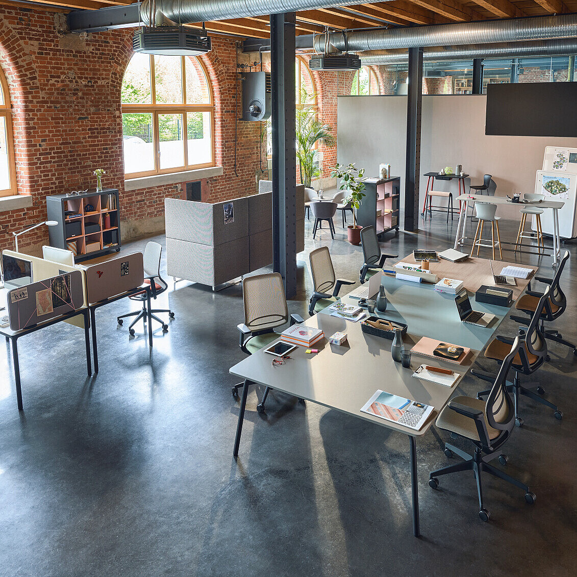 Open-plan office with multiple desks, chairs, computers, and large arched windows in a brick-walled room