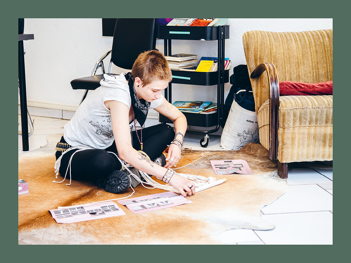 Person sitting cross-legged on a light brown animal hide rug, arranging pink papers with black-and-white images, near a beige armchair and a black chair in a room.