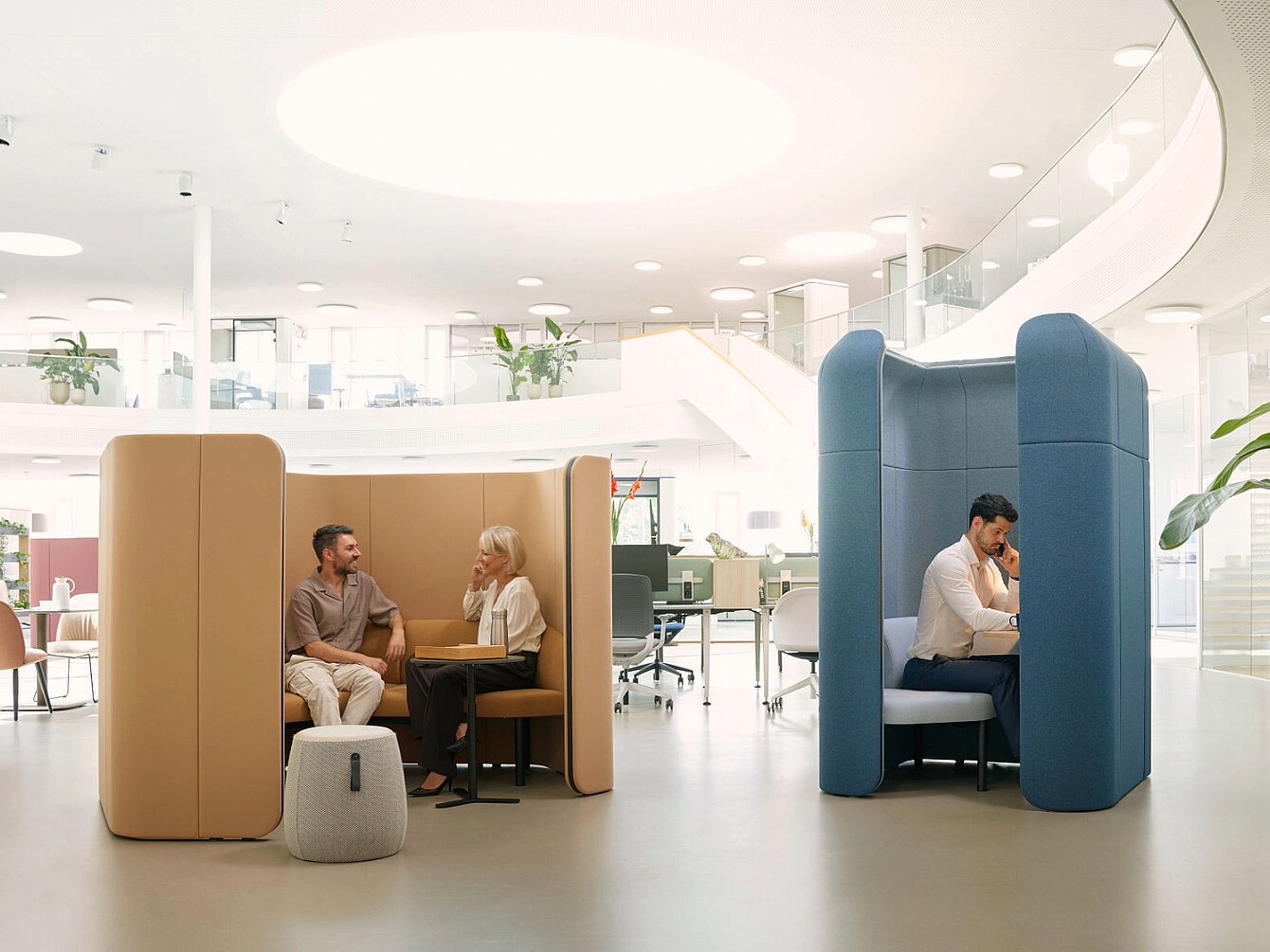 Zwei Personen sitzen in separaten se:hive Raumlösungen von Sedus, eine beige und eine blaue, in einem offenen, hellen Büro mit moderner Einrichtung.