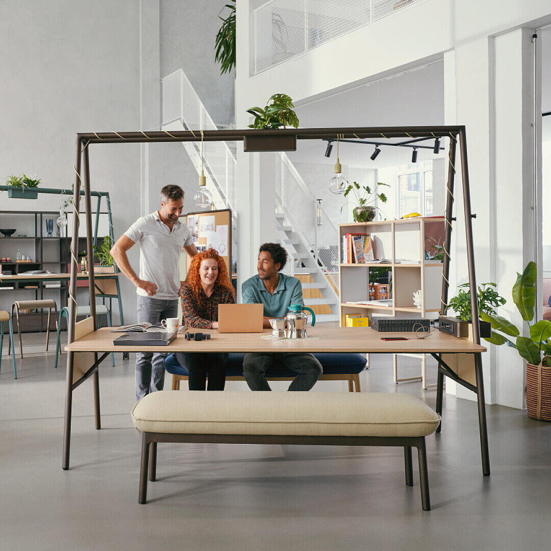 Three employees collaborating at a large wooden table in a bright, open office with natural light, plants, and modern furniture, illustrating a workplace focused on wellbeing and social spaces.
