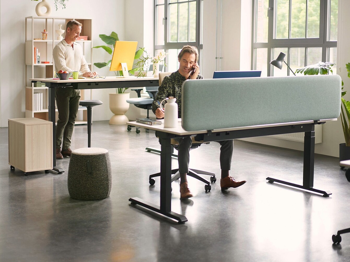 Two men working in a modern office with large windows one standing at a desk with a yellow screen and the other sitting behind a blue fabric desk divider talking on the phone