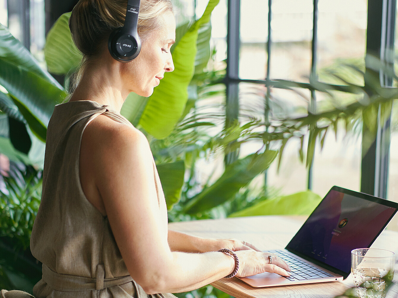 Person wearing headphones working on a laptop at a wooden table surrounded by large green plants in a Garden Work Café setting