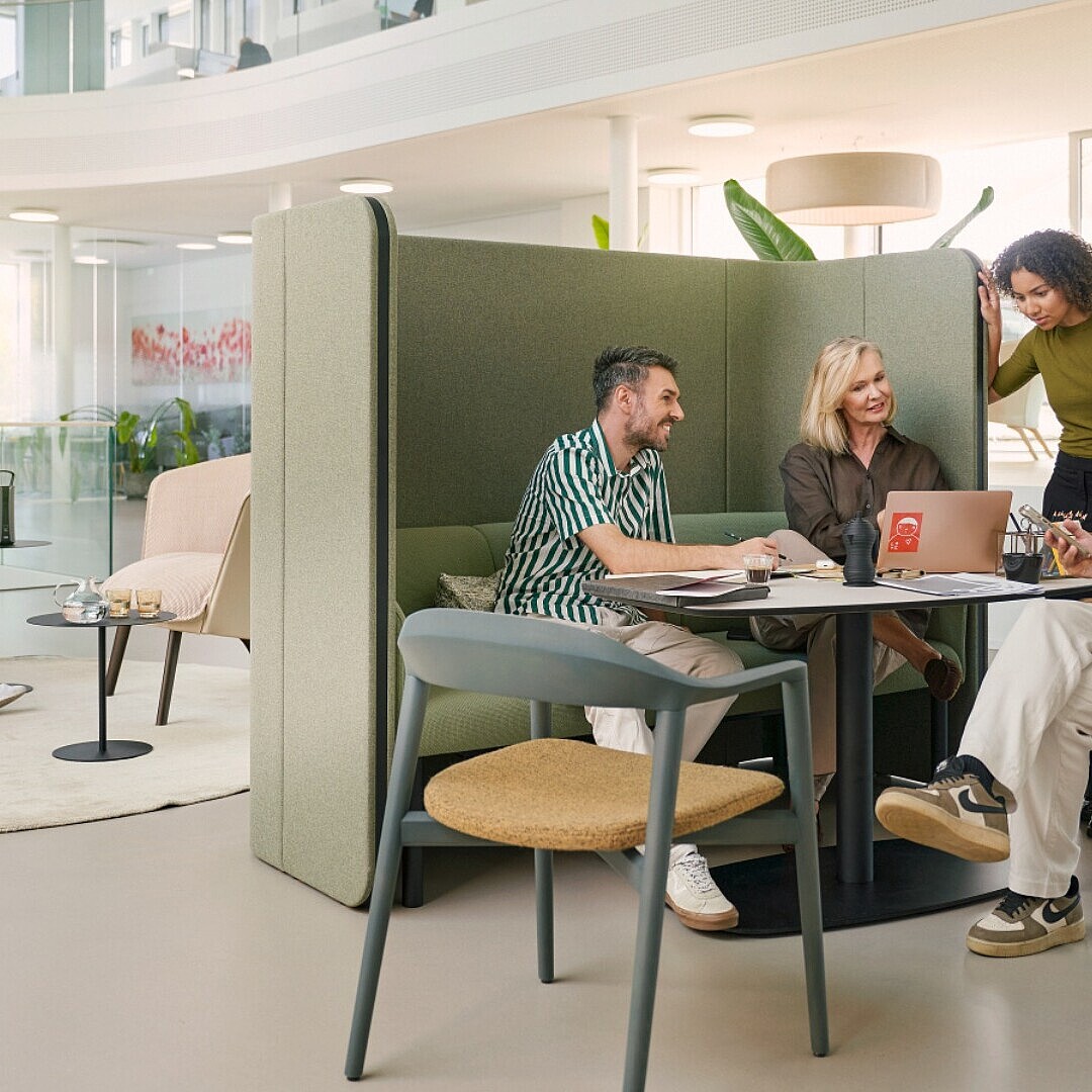 Five people sitting and standing in a modern open office with plants and large windows, three at a table with laptops and notebooks, two on armchairs with a small table between them