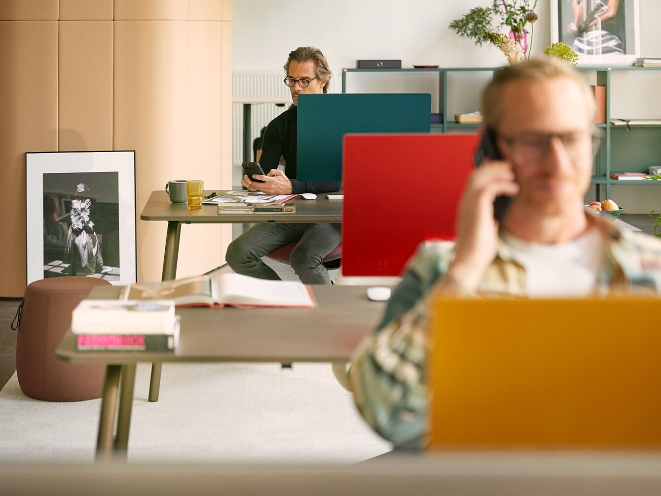 Zwei Mitarbeiter arbeiten an Schreibtischen mit farbigen Sichtschutzwänden, während eine Person im Vordergrund telefoniert.