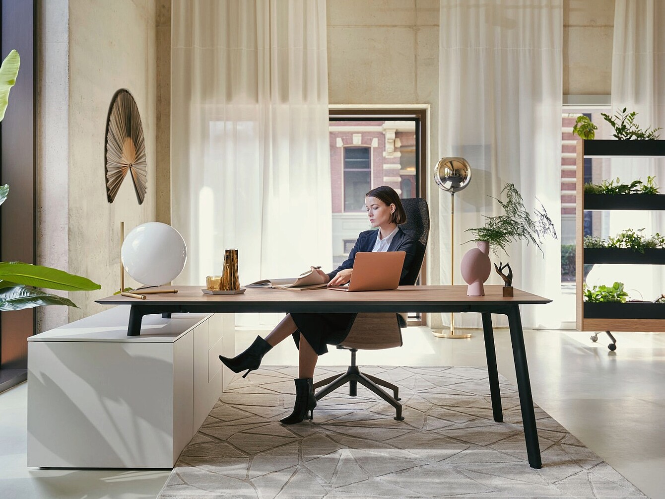 Woman sitting at a modern office desk with a laptop and notebook in a bright room with sheer curtains, plants, and decorative objects
