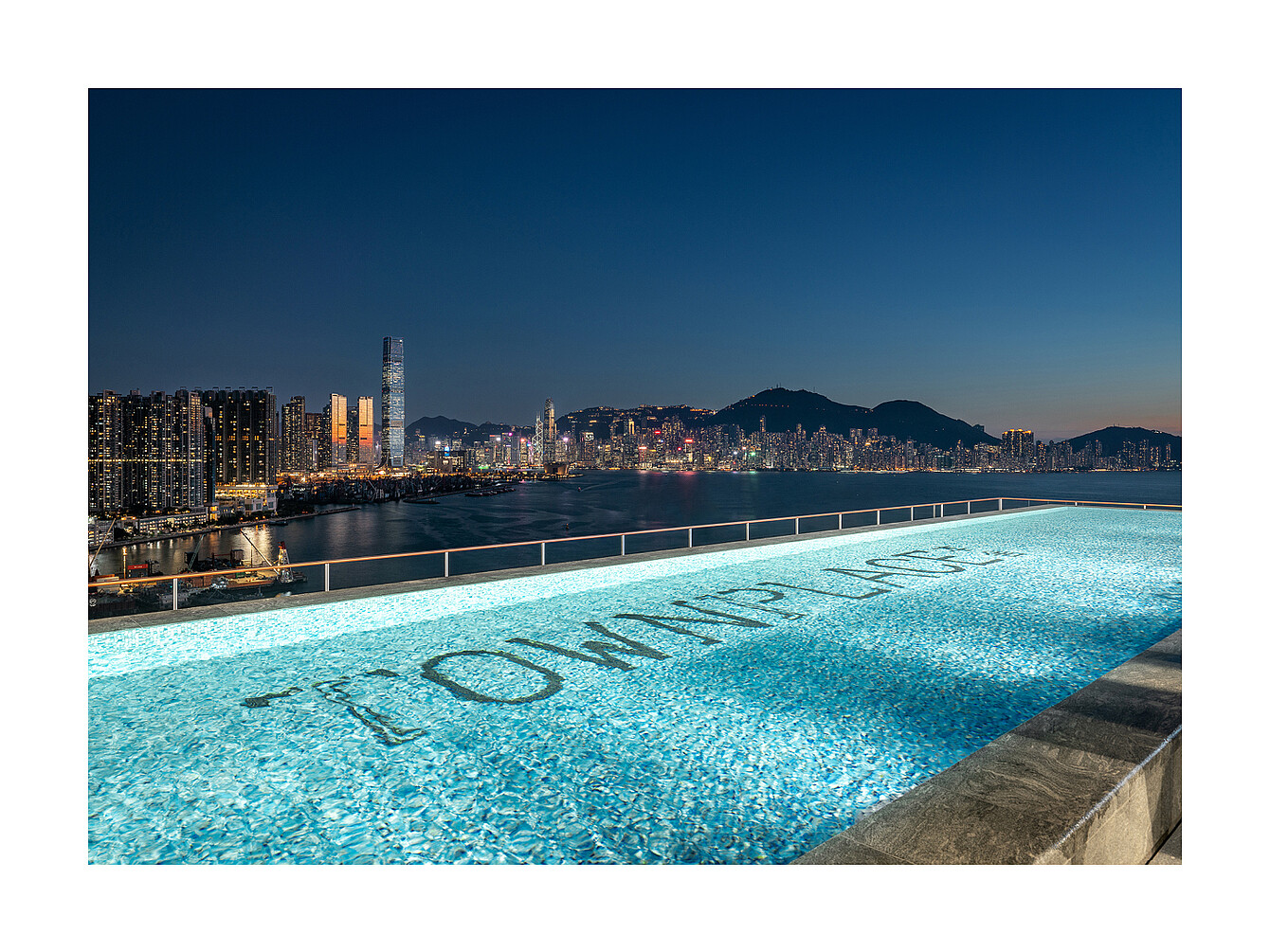 Rooftop infinity pool with the word 'TOMMY LIFE' visible on the pool floor overlooking a city skyline at dusk