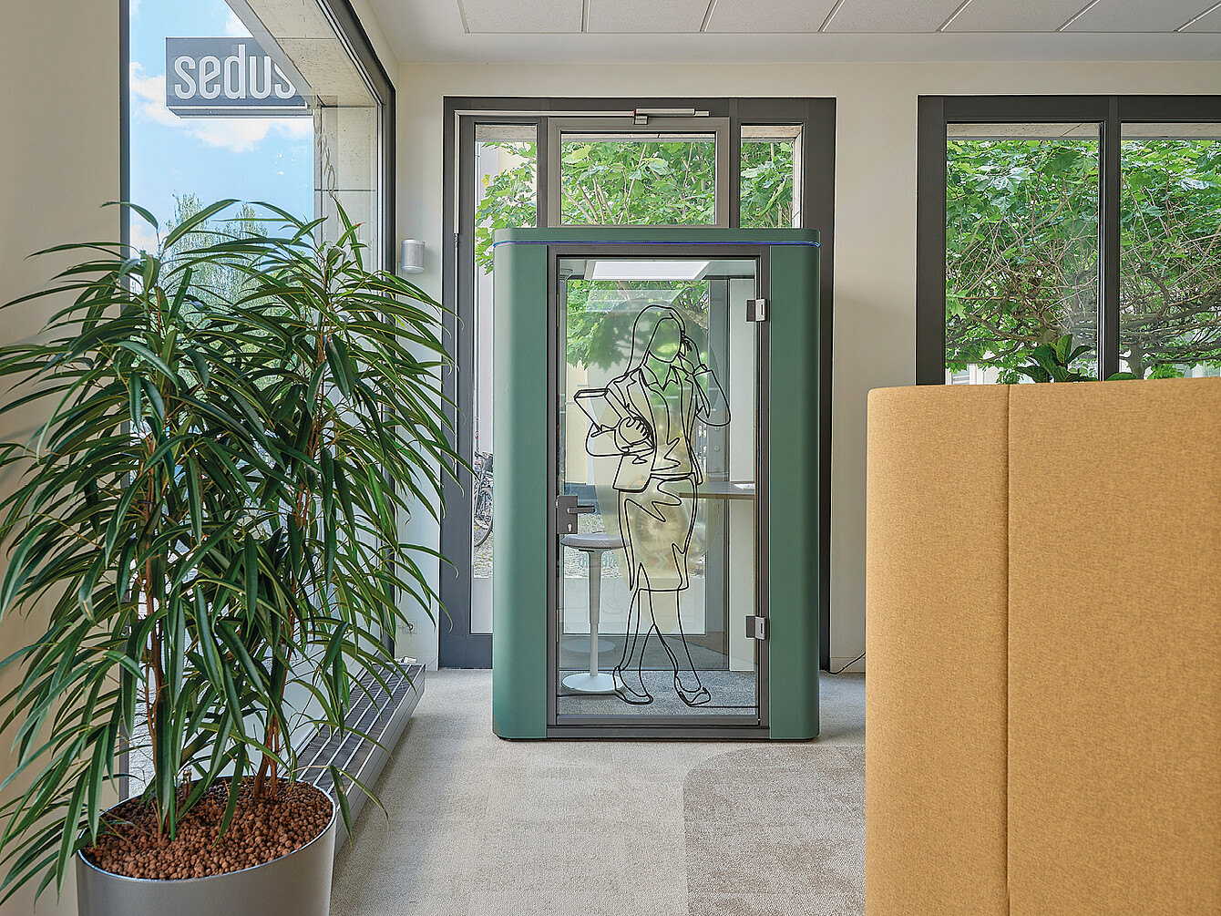 Small green soundproof booth with a glass door featuring a line drawing of a woman holding a phone, placed in a modern office space near large windows and a tall potted plant.