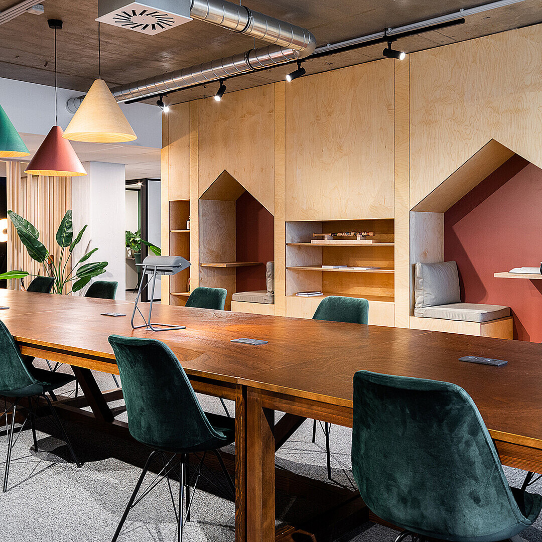 Open office space with a long wooden table surrounded by green upholstered chairs and built-in wooden seating niches along the wall under pendant lights