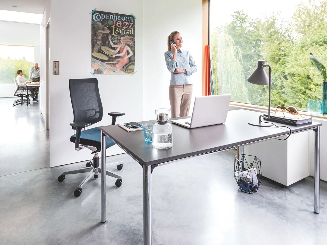 Modern office with a black mesh ergonomic chair, a desk holding a laptop, glass water bottle, lamp, and a woman standing by a large window talking on a phone