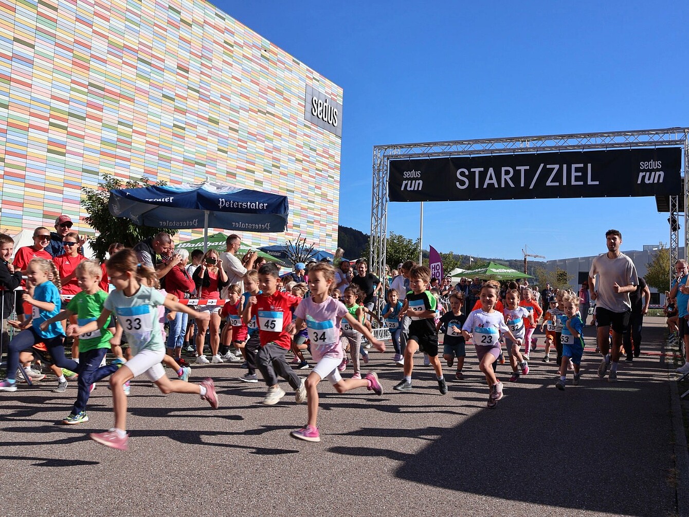 Group of children running at the start line of the Sedus run event under a banner reading 'START/ZIEL' with spectators and a building in the background