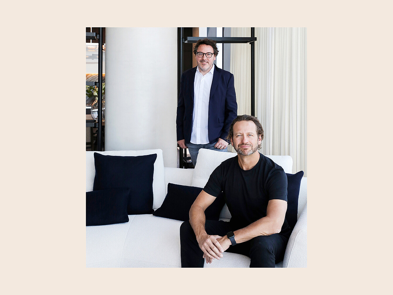 Two men in a modern interior with a white sofa and black cushions, one sitting and one standing behind the sofa.