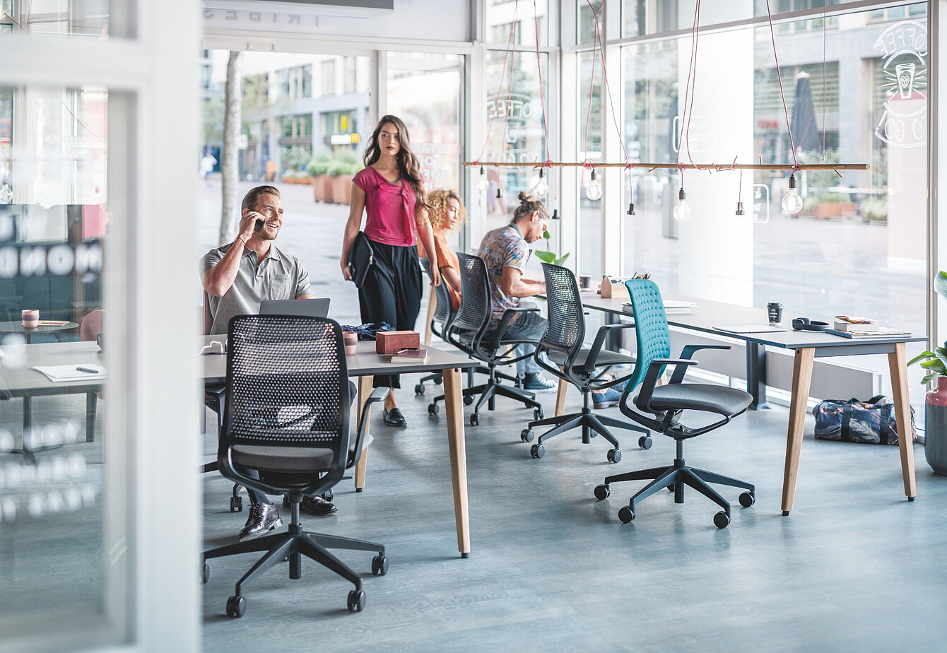 Three people in a modern office with large windows and mesh office chairs around a wooden table