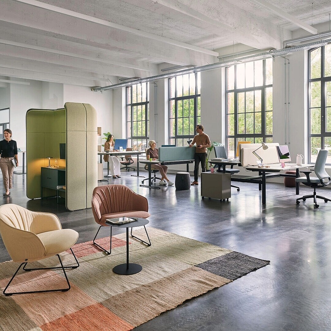Offenes Büro mit großen Fenstern, mehreren Schreibtischen, zwei schallisolierenden Sitzkabinen und zwei Sesseln auf Teppich