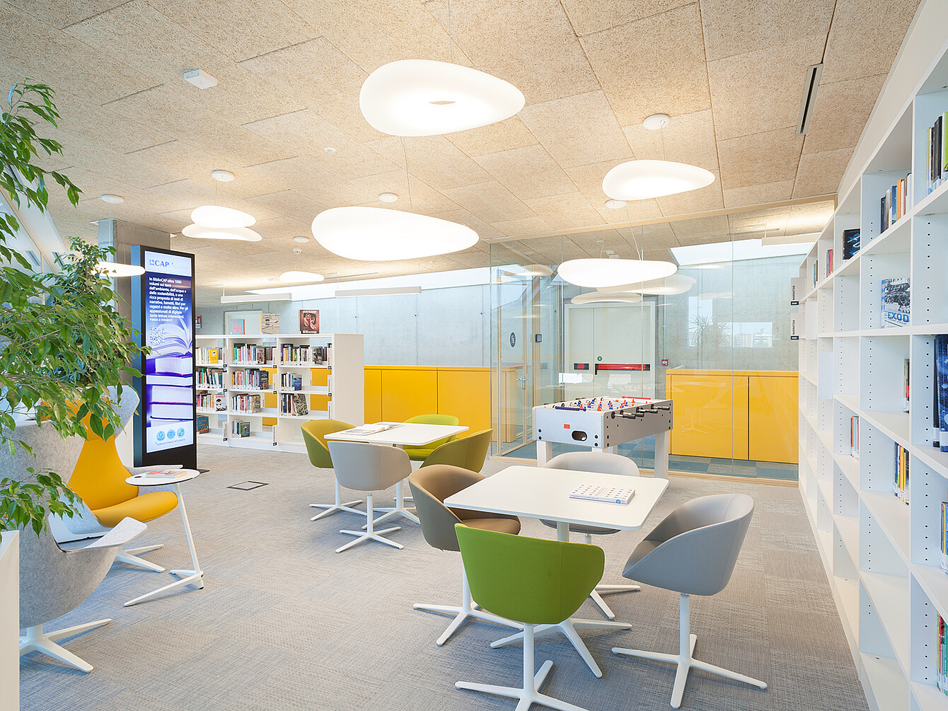Open office area with white tables surrounded by green and gray swivel chairs, white bookshelves, yellow storage units, a foosball table, and large ceiling lights