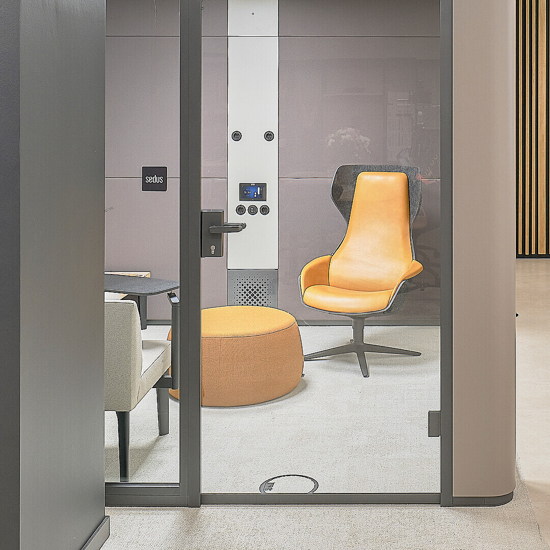 Modern office space with a glass-walled meeting room containing an orange swivel chair and round ottoman, adjacent to a lounge area with a green sofa, small round stool, and wooden slat wall with plants and coat rack.