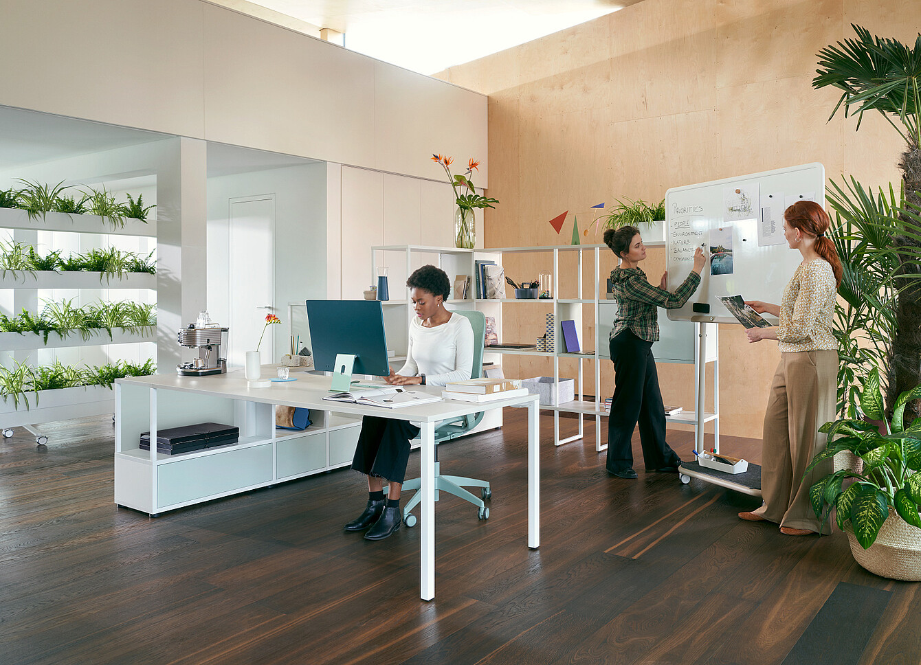 Three women in a modern office with plants one sitting at a desk working on a computer and two standing by a whiteboard discussing notes