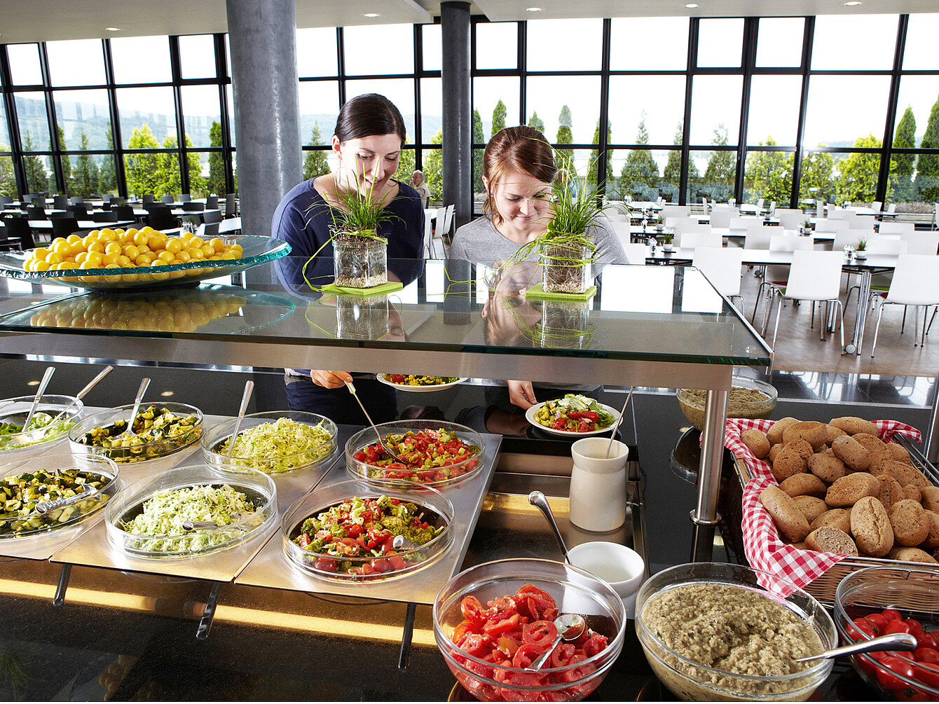 Two women selecting fresh salads and healthy food options at a sustainable catering buffet in a modern, bright workplace cafeteria