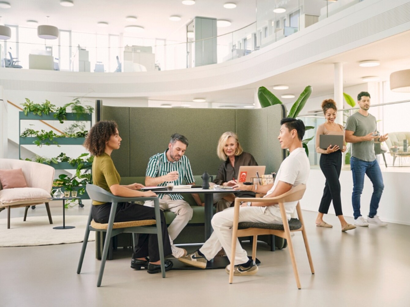 Four people sit around a round table in an open office while two others walk by in the background