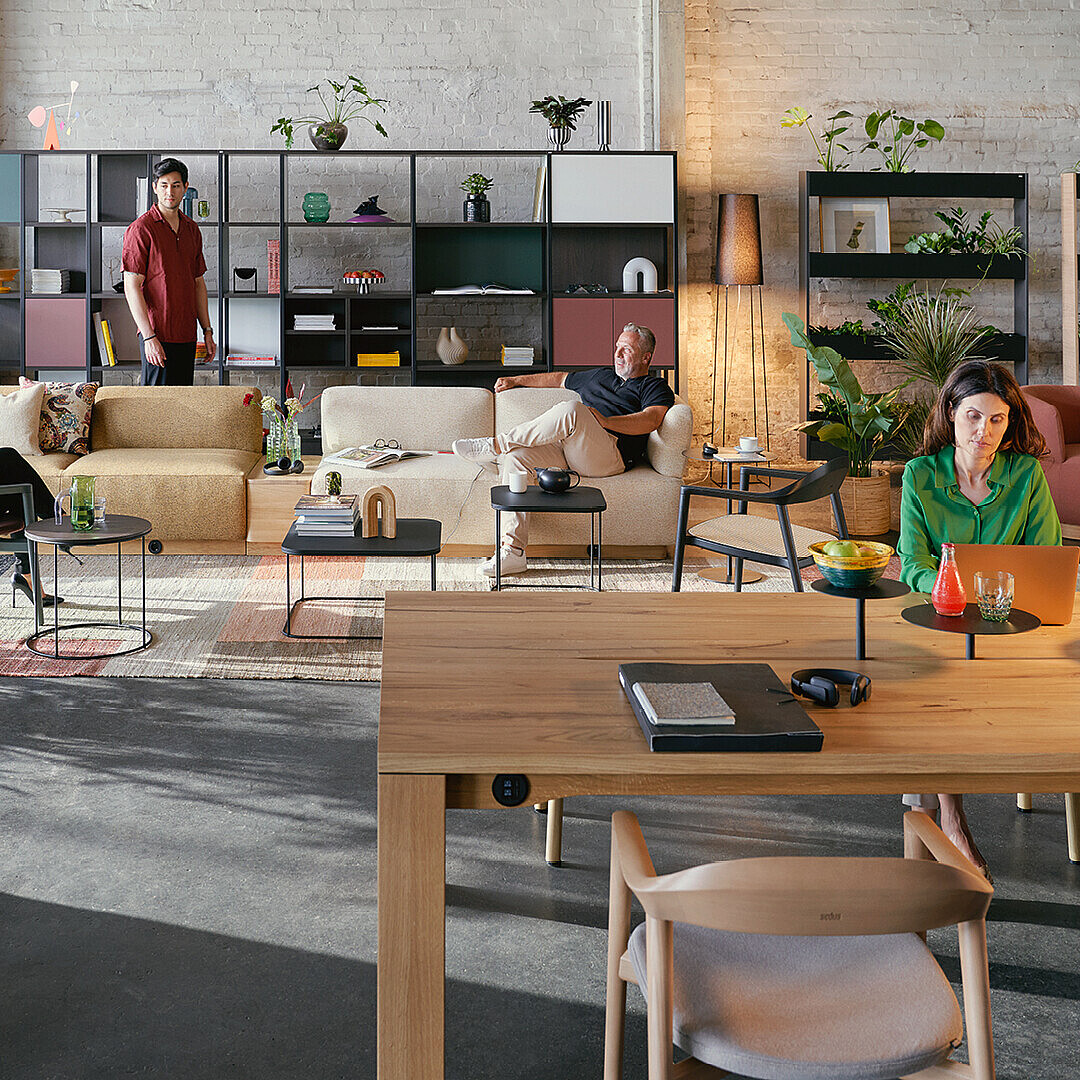 Modern hybrid ductile commons workspace with diverse people using sofas, chairs, and a large wooden table surrounded by plants and shelves, promoting flexibility and community.