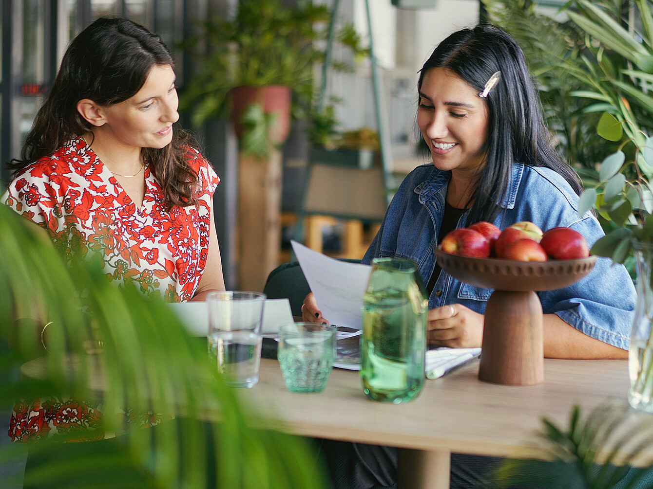 Two women engaged in a friendly discussion over documents at a wooden table in a bright office space with plants and a bowl of apples, illustrating intentional office visits for meaningful interaction and support.