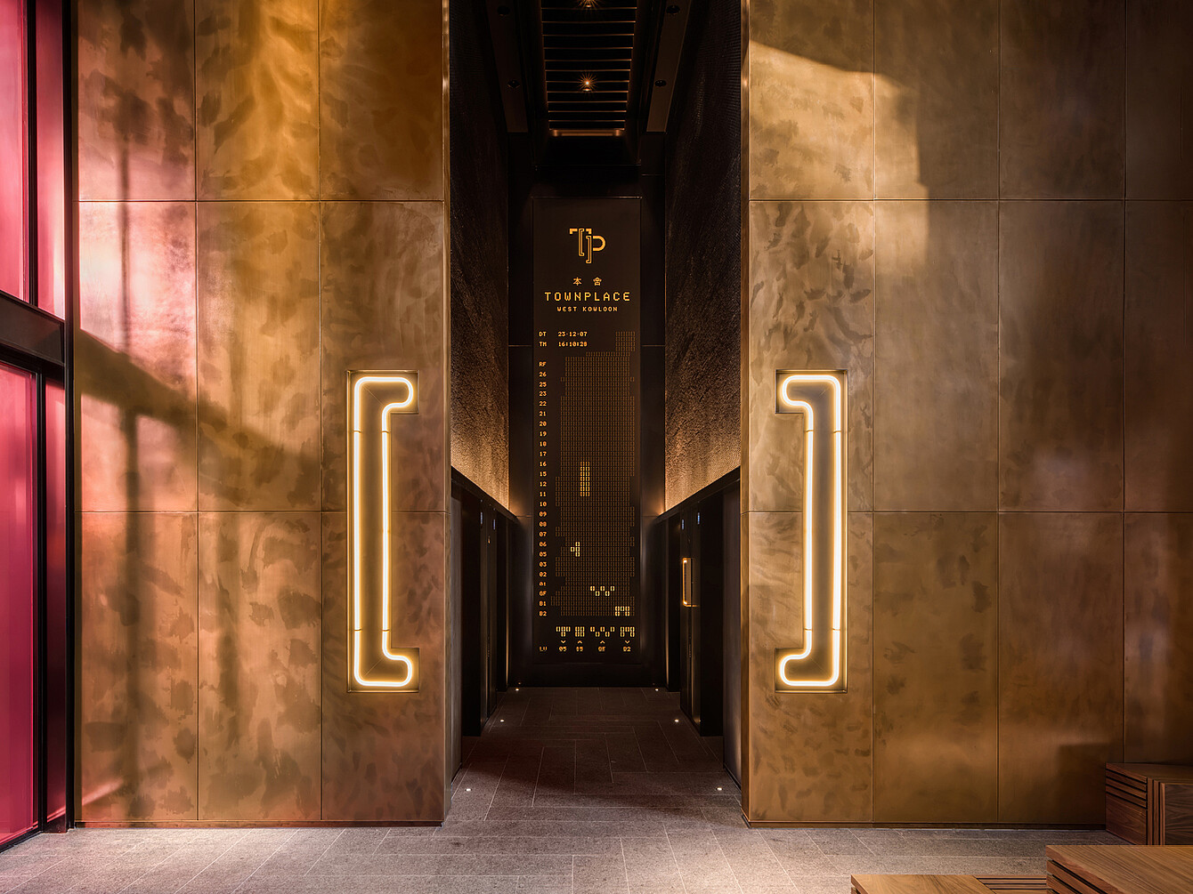 Narrow corridor with bronze-colored walls and illuminated vertical handles on each side leading to a dark entrance with a sign reading 'TP TOWNPLACE'