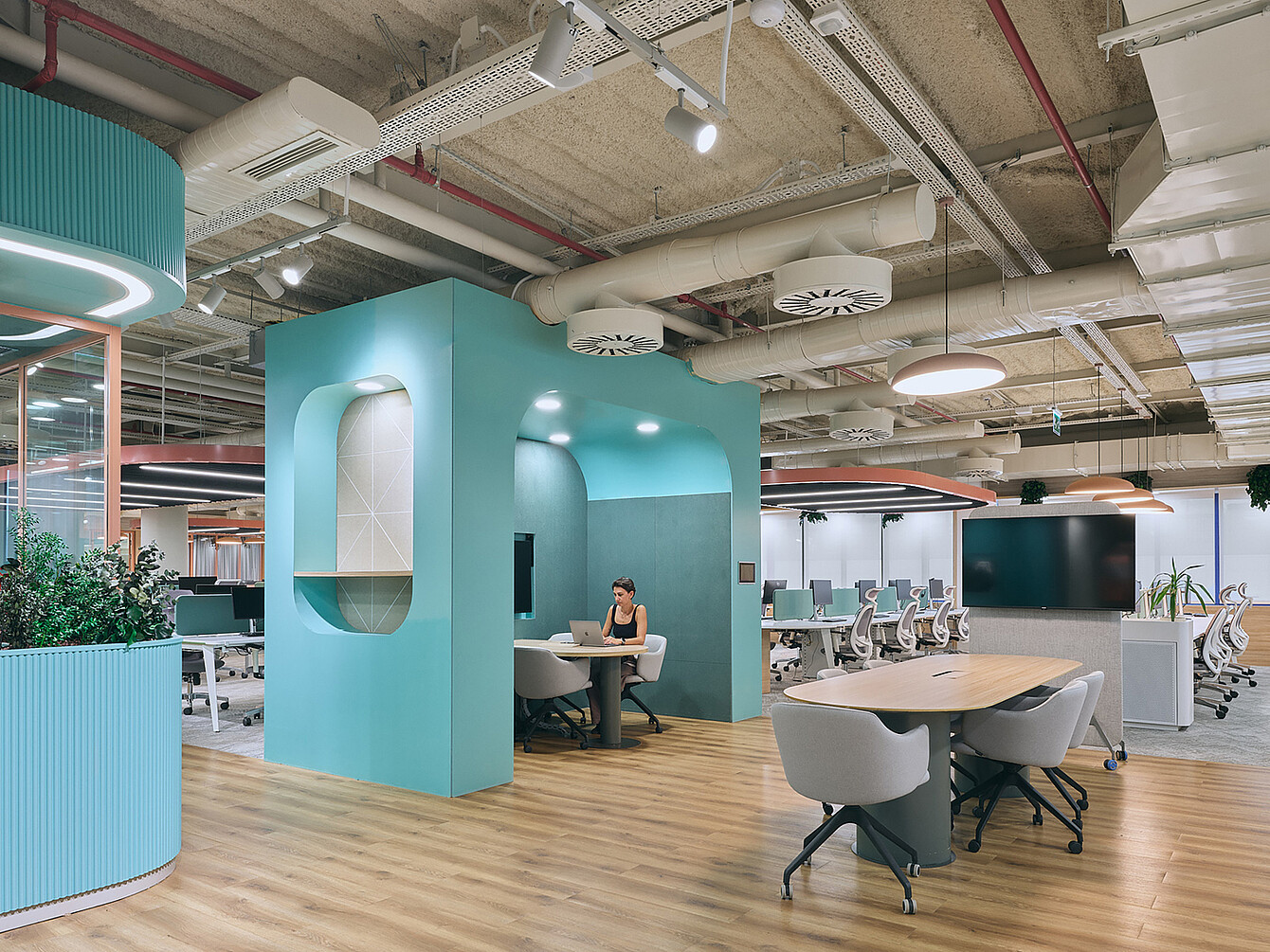 Open-plan office space with light wood flooring, a turquoise enclosed meeting pod with a person working on a laptop inside, multiple white office chairs, desks with computer monitors, and exposed ceiling pipes and ducts