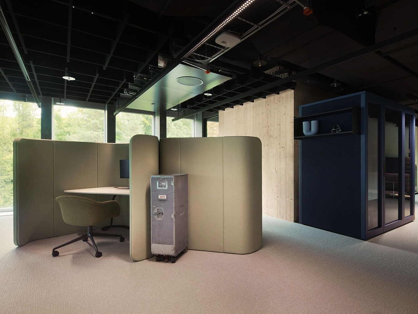 Arbeitsbereich mit beige-farbenen, hohen Trennwänden, einem grauen Bürostuhl auf Rollen und einem grauen Metallkoffer auf dem Boden, im Novu Campus am Züricher Flughafen.