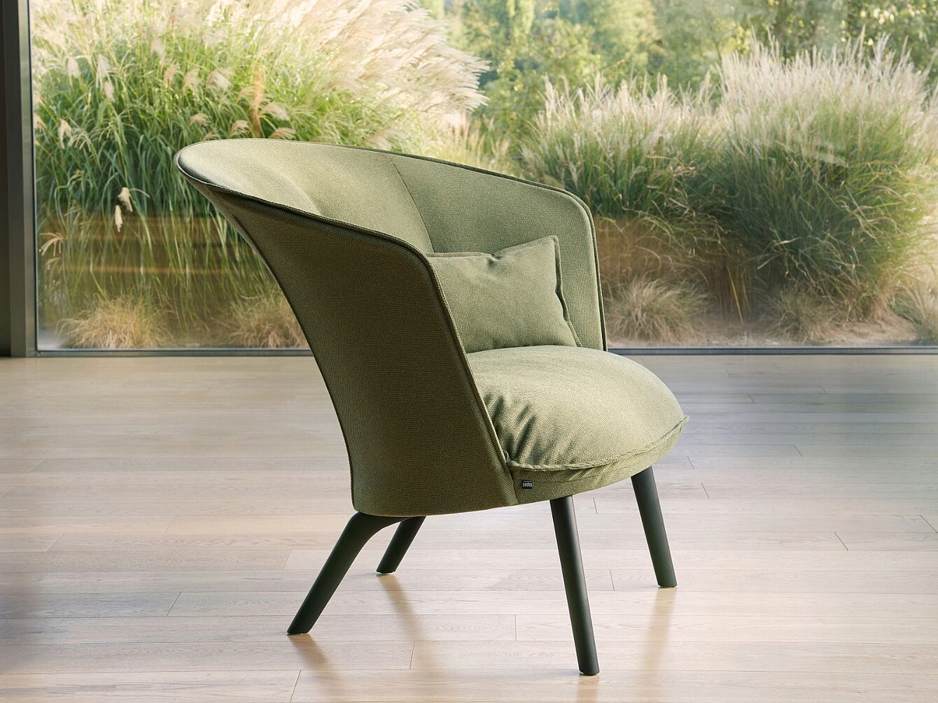 Modern armchair with green fabric upholstery and small cushion in front of large glass window showing garden plants