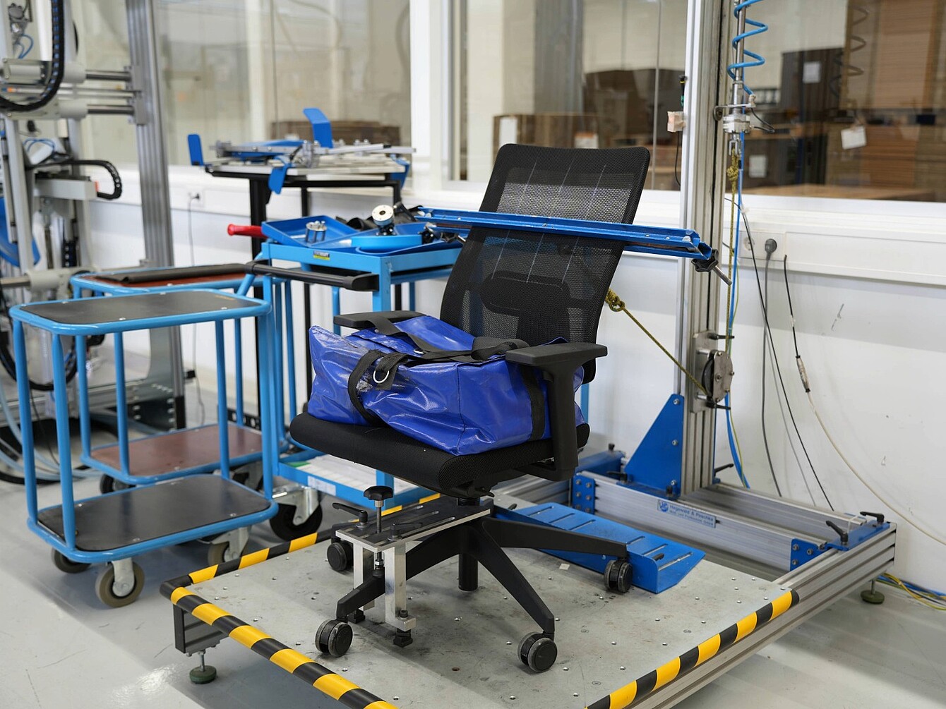 Office chair secured in a blue metal testing frame inside a laboratory with equipment and carts in the background