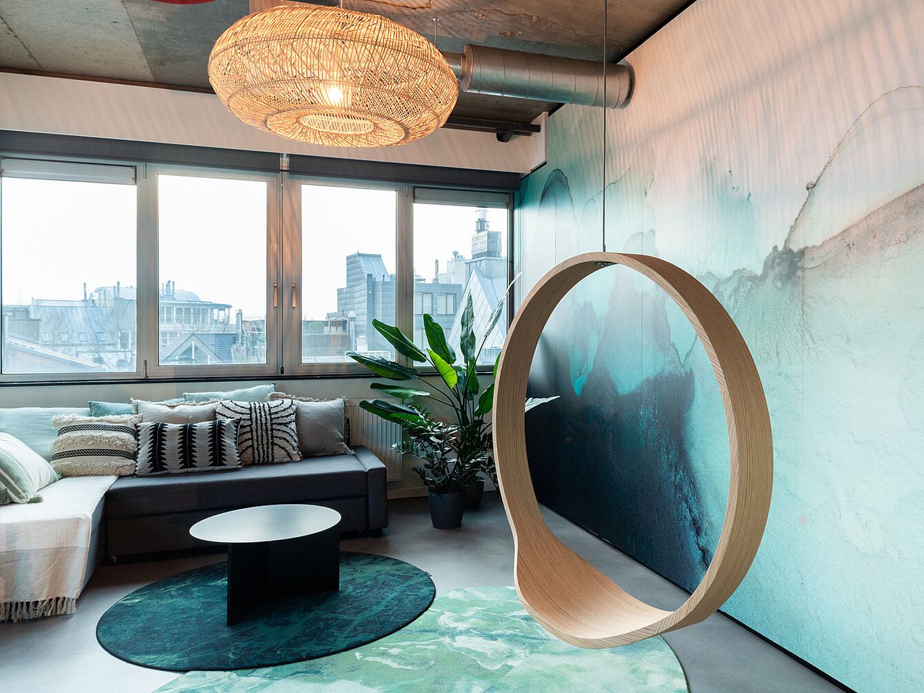 Modern office lounge with a gray sectional sofa, patterned cushions, a round black coffee table on a green rug, a large plant, a circular wooden hanging chair, and a large window showing city buildings