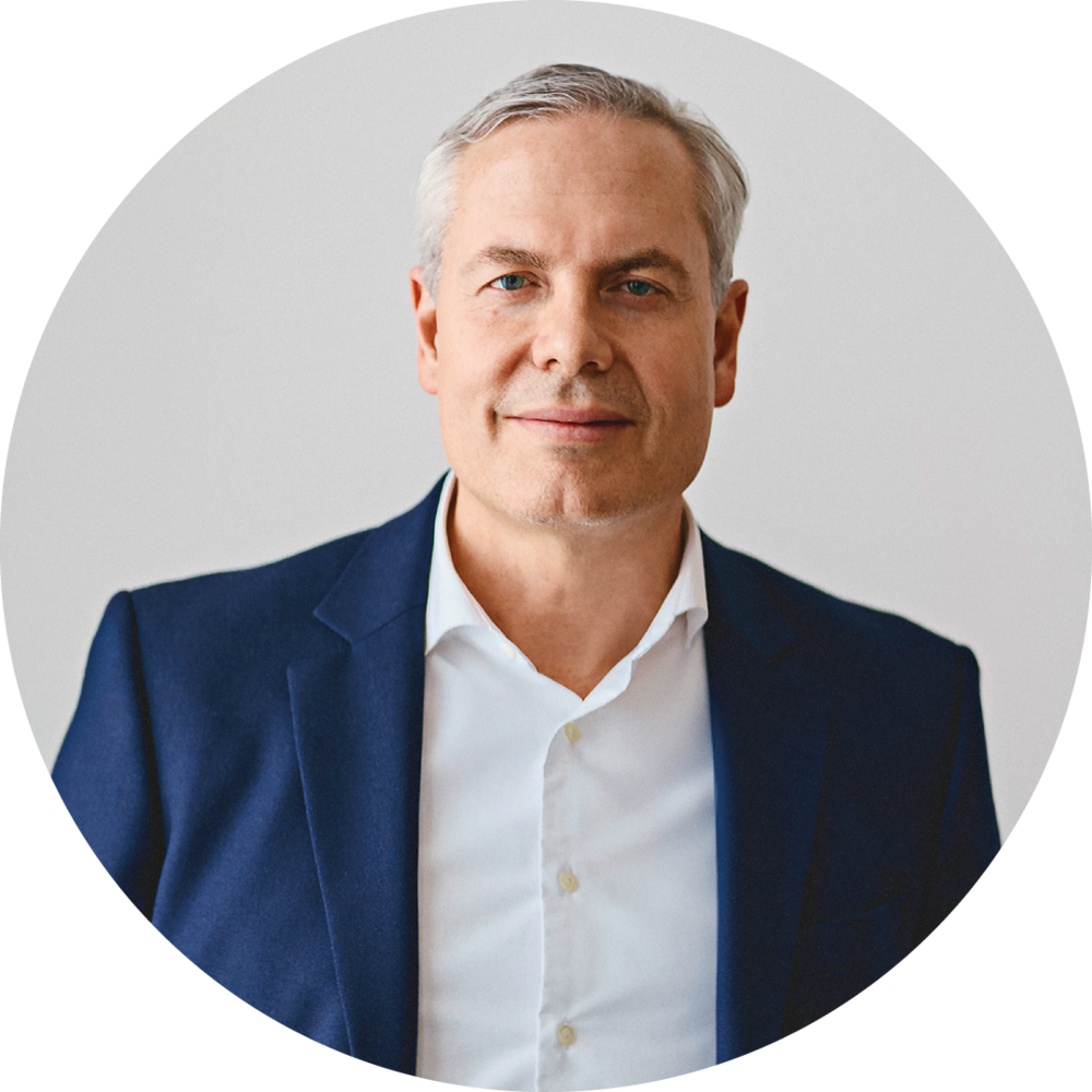 Middle-aged man (Ernst Holzapfel) with short gray hair wearing a white shirt and dark blazer against a plain light background