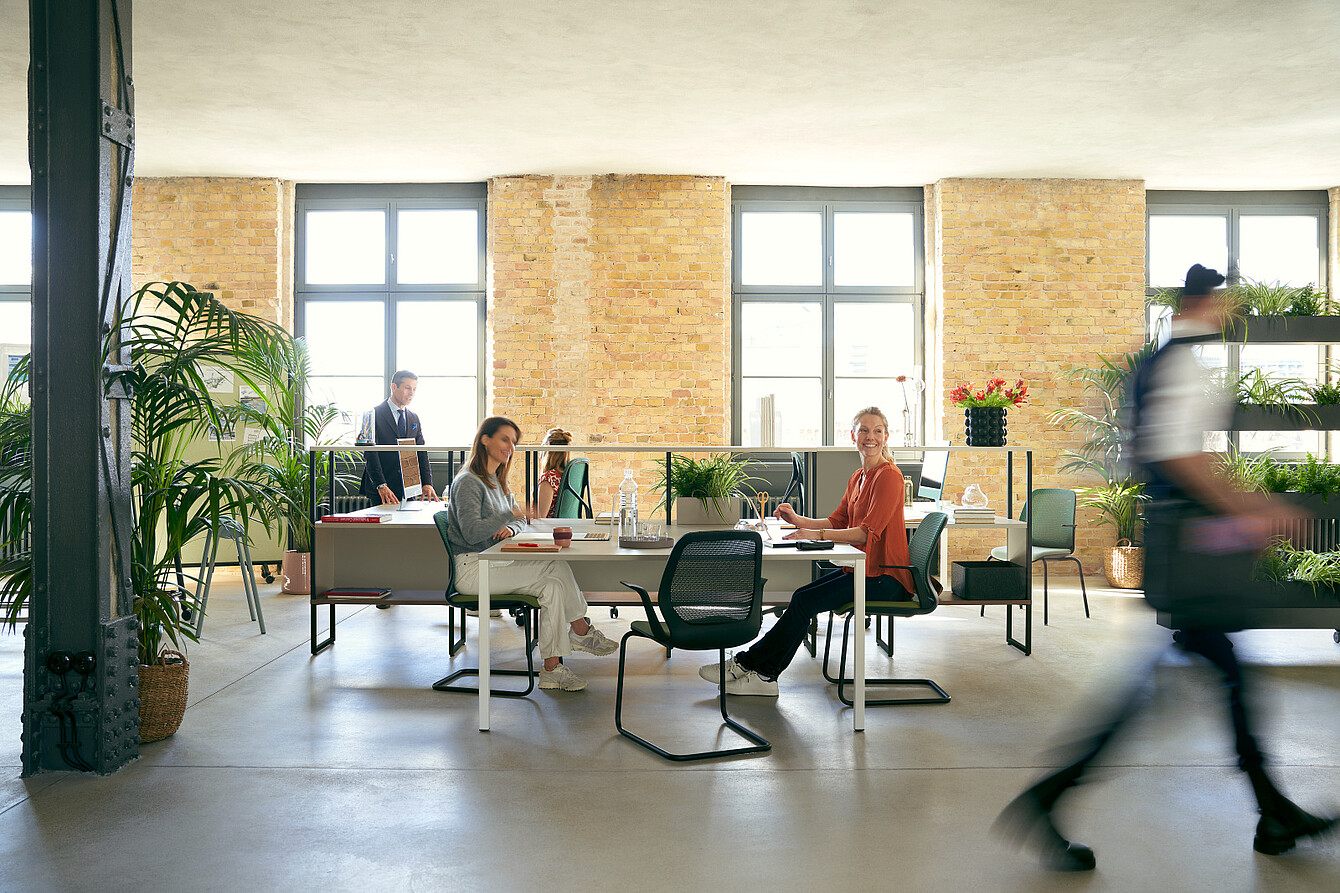 Two women sitting at desks in a spacious office with large windows and exposed brick walls while a man walks past in the foreground and another man stands near the windows