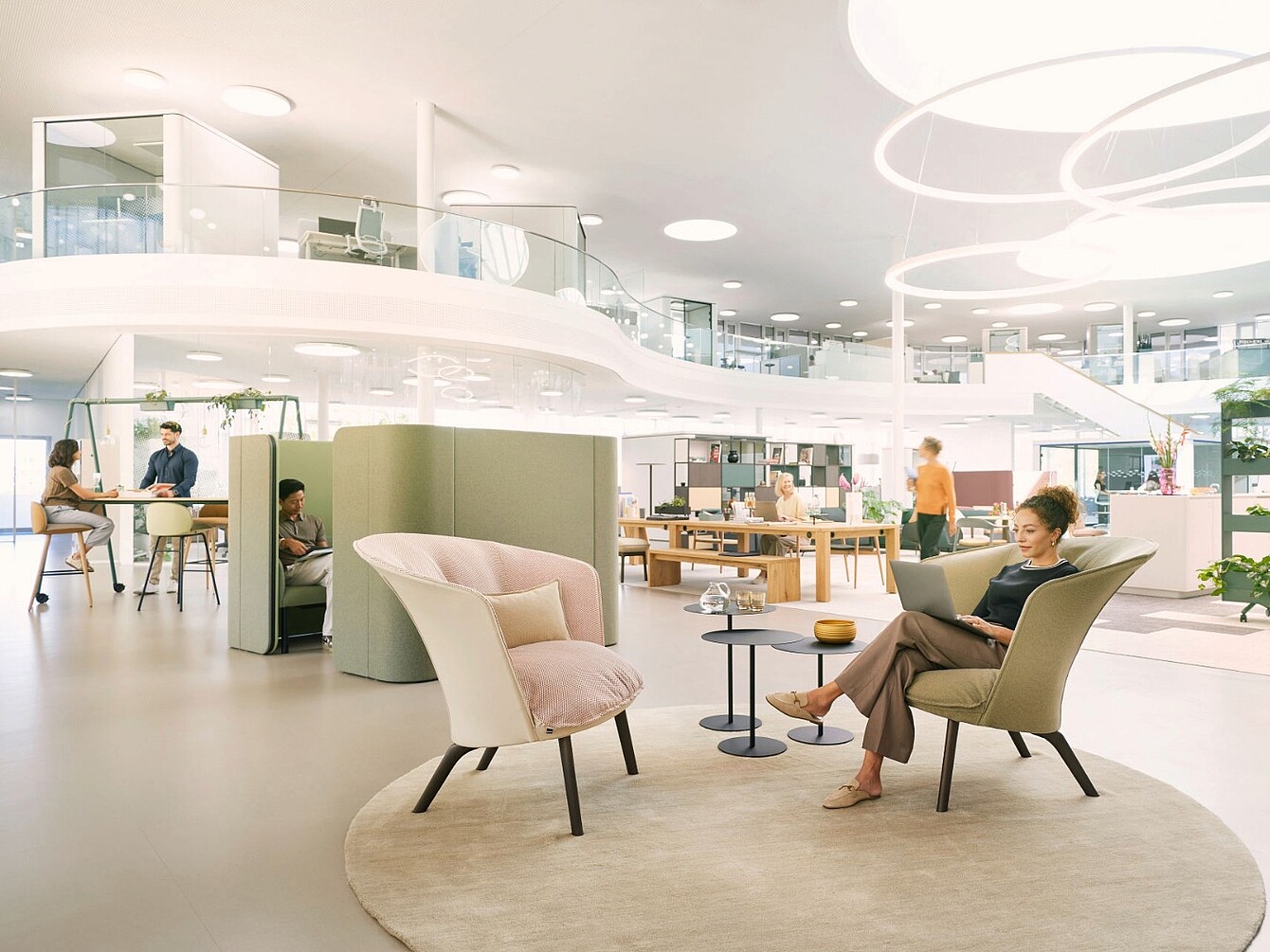 Modern open office space with a woman sitting in a beige armchair reading a book, a small round table beside her, and other people working or conversing in the background
