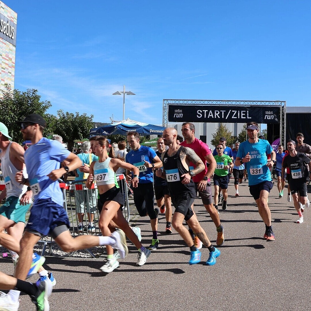 Group of runners starting a race outdoors near a building with a 'Sedus' sign and a black start/finish arch